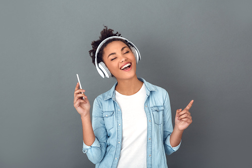 Young african female student isolated on grey wall listening to popular music holding smartphone joyful fun