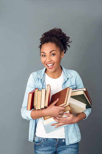 Young african female student isolated on grey wall with a messed pile of books looking camera playful