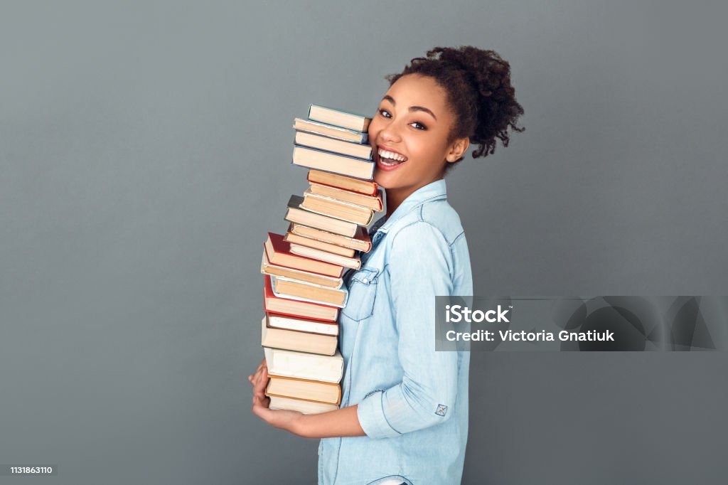Young african woman isolated on grey wall studio casual daily lifestyle holding books profile Young african female student isolated on grey wall holding a pile of books standing profile looking camera laughing Book Stock Photo