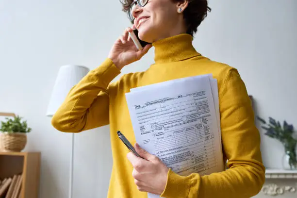 Close-up of positive young woman in glasses standing in living room and holding individual income tax return form while talking by mobile phone