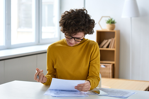 Serious concentrated young woman in glasses sitting at table and analyzing tax documents in home office