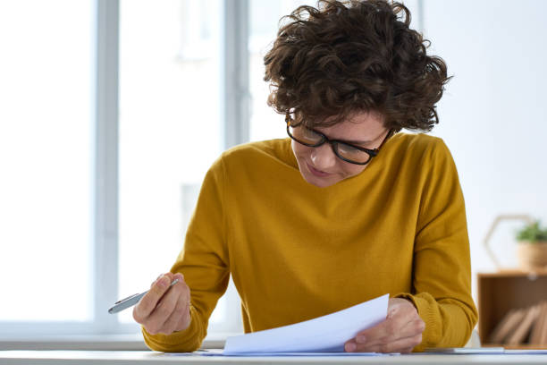 senhora nova ocupada séria com o cabelo curly curto que senta-se na mesa e no documento de enchimento ao preparar papéis de imposto - form filling - fotografias e filmes do acervo