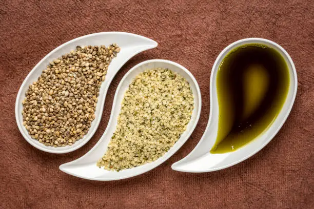 dry hemp seeds, hearts and oil in small teardrop bowls against brown textured bark paper