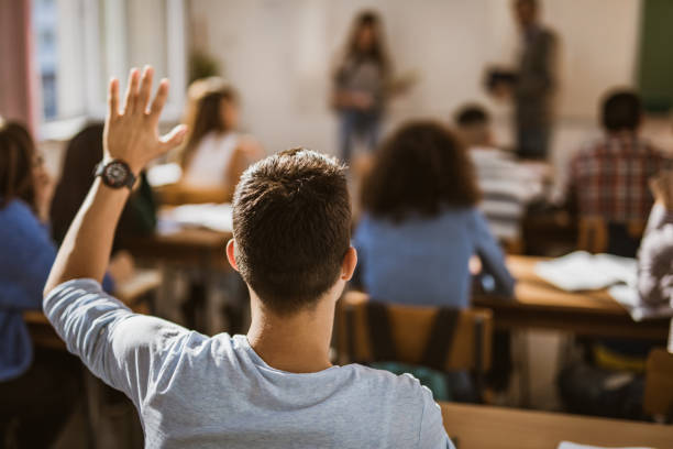 Back view of male student raising hand on a class. Rear view of a male student raising his hand to answer the question on a class at school. hand raised classroom student high school student stock pictures, royalty-free photos & images
