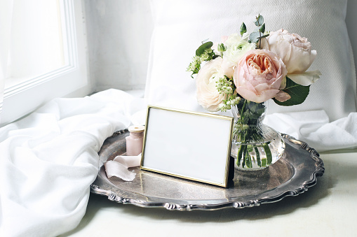 Spring, summer still life scene. Blank golden photo frame old silver tray at windowsill. Vintage feminine styled photo. Floral composition, bouquet of pink English roses, Ranunculus and eucalyptus.