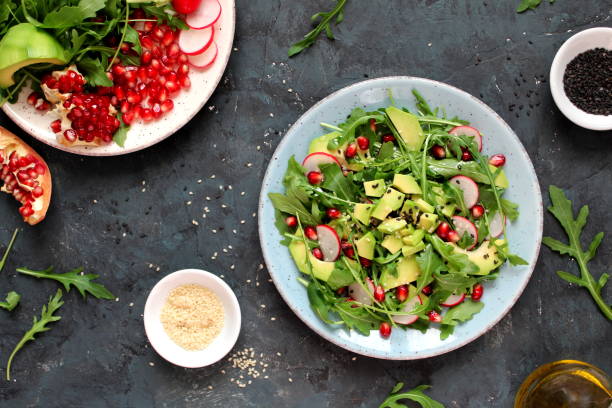 Ingedient for making salad on dark background. Vegetable salad in plate, avocado, arugula, pomegranate, radish and sesame.
