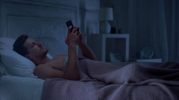 istock Sleepy male lying on bed and using cellphone, setting alarm for morning wake 1131889100