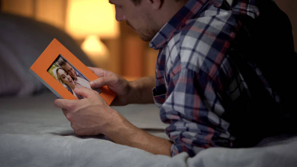 mladý muž v zoufalství ležící na posteli a dívá se na fotografii s přítelkyní, smutek - potíže ve vztazích fotky - stock snímky, obrázky a fotky