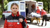Paramedic posing for camera, ambulance crew transporting patient on background