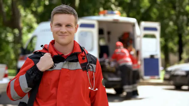 Male doctor smiling into camera, ambulance crew working, blurred on background