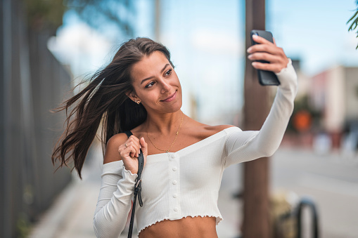 Smiling beautiful woman taking selfie on mobile phone. Young female with tousled hair is wearing crop top. She is enjoying in city