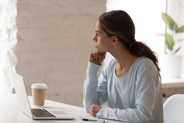 Concerned female sitting at workplace thinking looking away Mixed race concerned female sitting at workplace desk opposite computer holding head with hand thinking looking out the window having problems and doubts in making decision searching solution concept Distracted stock pictures, royalty-free photos & images