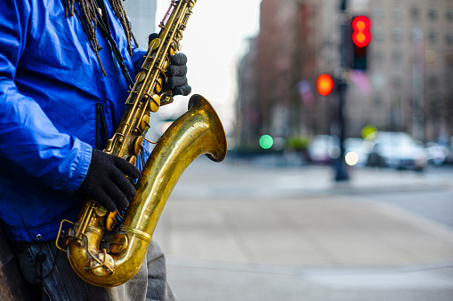 A horizontal cropped view of an unrecognizable street saxophone player in a blue jacket and dreadlocks with colorful bokeh lights in the background.