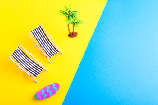 Miniature beach with deck chairs, palms and sufrboard on the colorful backgeound. Yellow and blue. Sand and ocean. Tropical resort and vacation concept