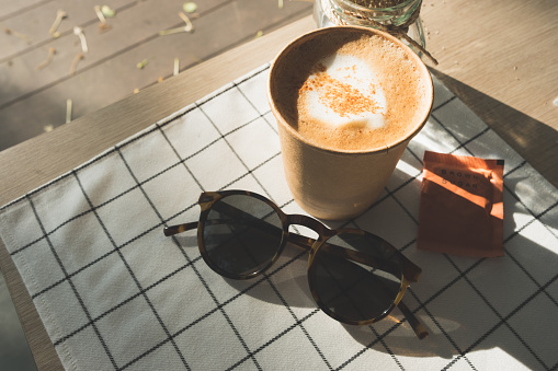 cappuccino in take away coffee cup on table cloth with dry flower,sunglasses on wood table with sunlight hard shadow at window in evening.food and drink lifestyle leisure concept