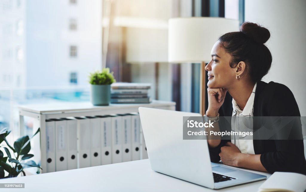 Dreams will get you far Shot of a young businesswoman using a laptop at her desk in a modern office and looking thoughtful Looking Through Window Stock Photo