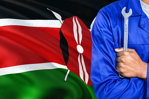 Kenyan Mechanic in blue uniform is holding wrench against waving Kenya flag background. Crossed arms technician.