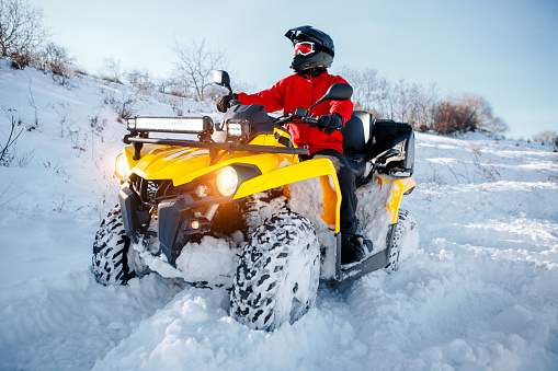 Young man driver in red warm winter clothes and black helmet on the ATV 4wd quad bike stand in heavy snow with deep wheel track. Moto winter sports