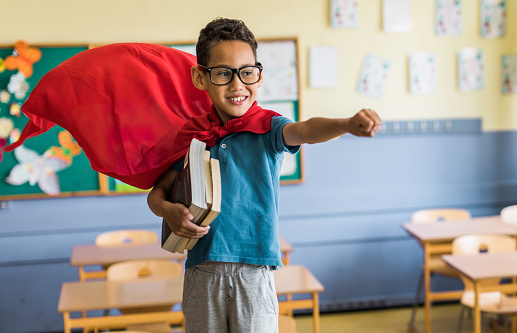 Playful African American elementary student having fun while pretending to be a superman in the classroom.