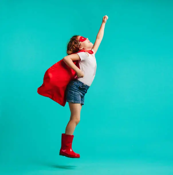 Adorable girl in red superhero costume and mask jumping with a arm raised. Girl in red cape, gumboot and mask fantasizing that she is superhero over blue background.