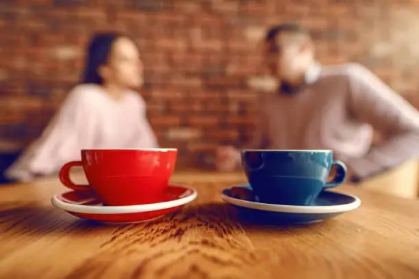 Photo of Close up of two cups of coffee on desk. In background blurred couple flirting.