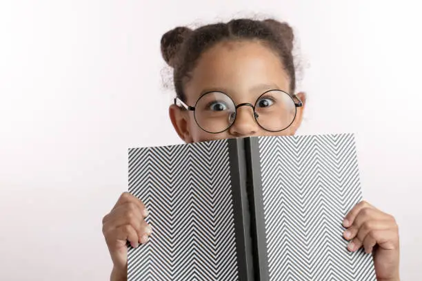 Photo of clever little girl with hairbunds and round glasses hiding behind the notepad