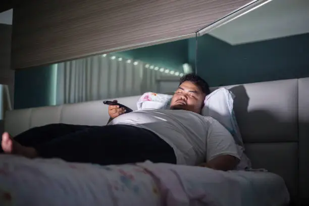 Picture of young fat man holding a remote TV while sleeping on the bed