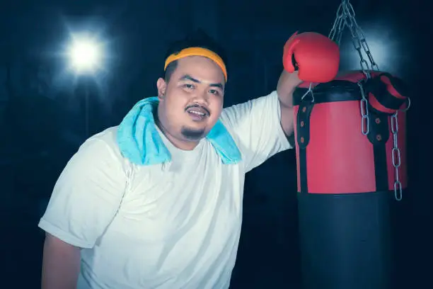 Picture of an obese man leaning on a bag boxing after boxing exercises in the dark room