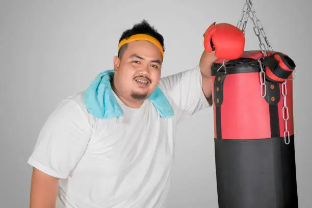 Picture of overweight man smiling at the camera while leaning on a bag boxing, isolated on white background