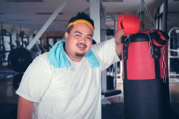 Picture of overweight man smiling at the camera while leaning on a boxing bag. Shot in the gym center