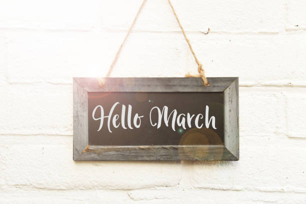 Hello March hand lettering on hanging sign board in spring sunlight Handwritten text hello March on hanging black chalkboard againt white outdoor wall march month photos stock pictures, royalty-free photos & images