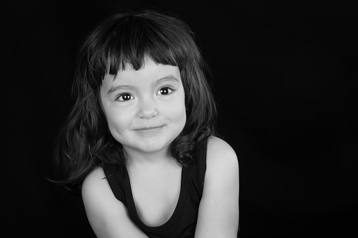 Black and white studio portrait of a beautiful toddler girl with black hair and brown eyes against a black backdrop