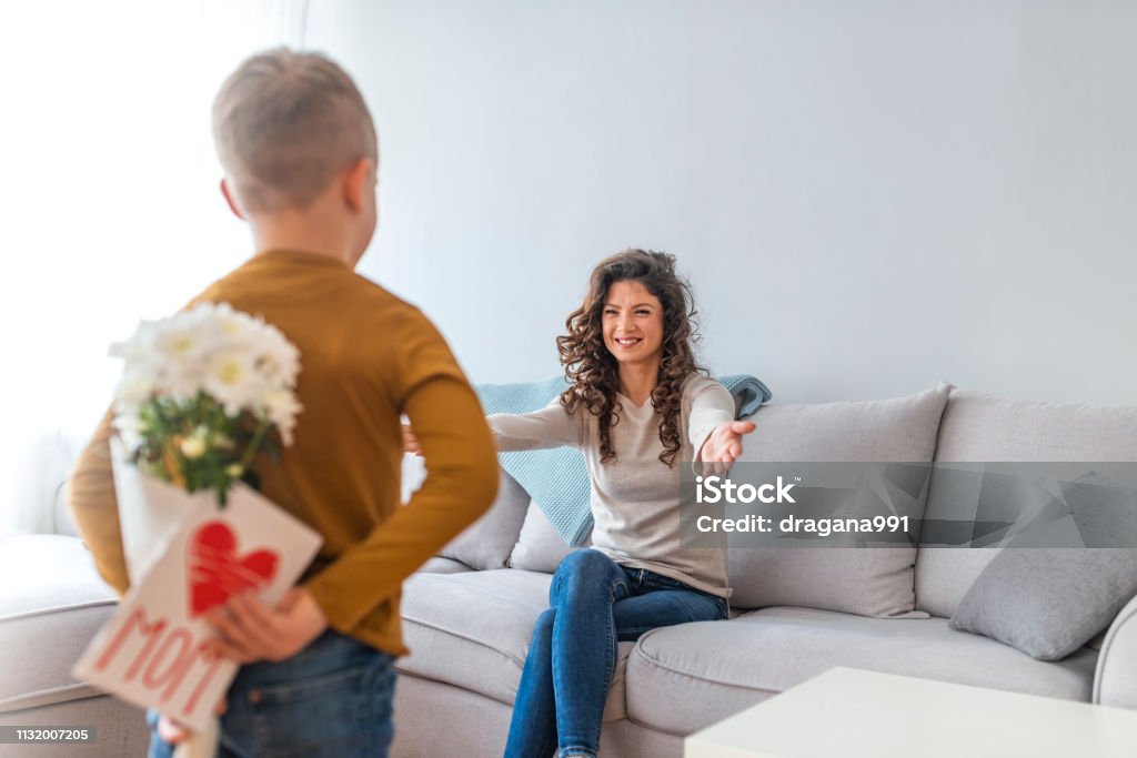 Mother's day concept Rear view of lad with bunch of beautiful flowers behind back preparing nice surprise for his mother. Cute boy offering flowers and card to his mother in the living room Mother's Day Stock Photo