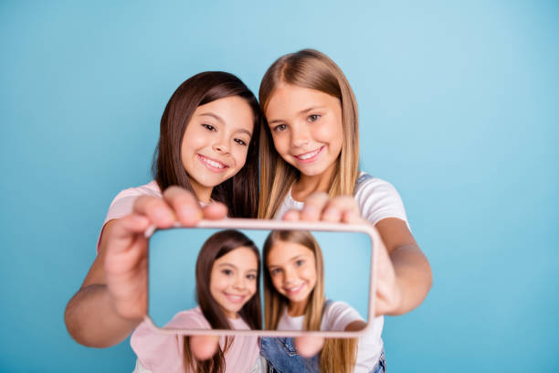 feche acima da foto dois pouco ela sua loira morena meninas longo cabelo bonito telefone fazer tomar selfies falar dizer falar millennials vestindo casual jeans denim t-shirts isoladas azul fundo brilhante - pre adolescent child little girls friendship child - fotografias e filmes do acervo