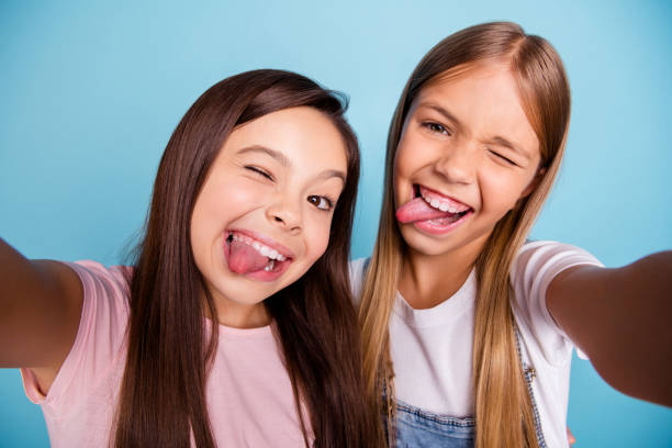 cerrar foto dos muy poco edad niñas vacaciones divirtiéndose lengua funky fuera de la boca hacen tomar selfies largo cabello bonito uso casual jeans denim camisetas aisladas en fondo azul brillante - video conference camera fotografías e imágenes de stock