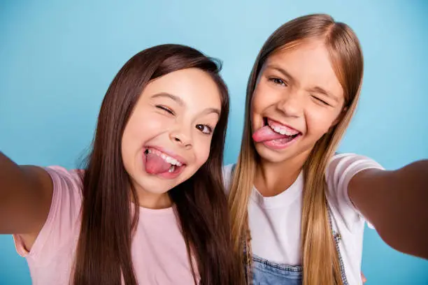 Close up photo two pretty little age girls holiday having fun funky tongue out of mouth make take selfies long pretty hair wearing casual jeans denim t-shirts isolated on blue bright background.