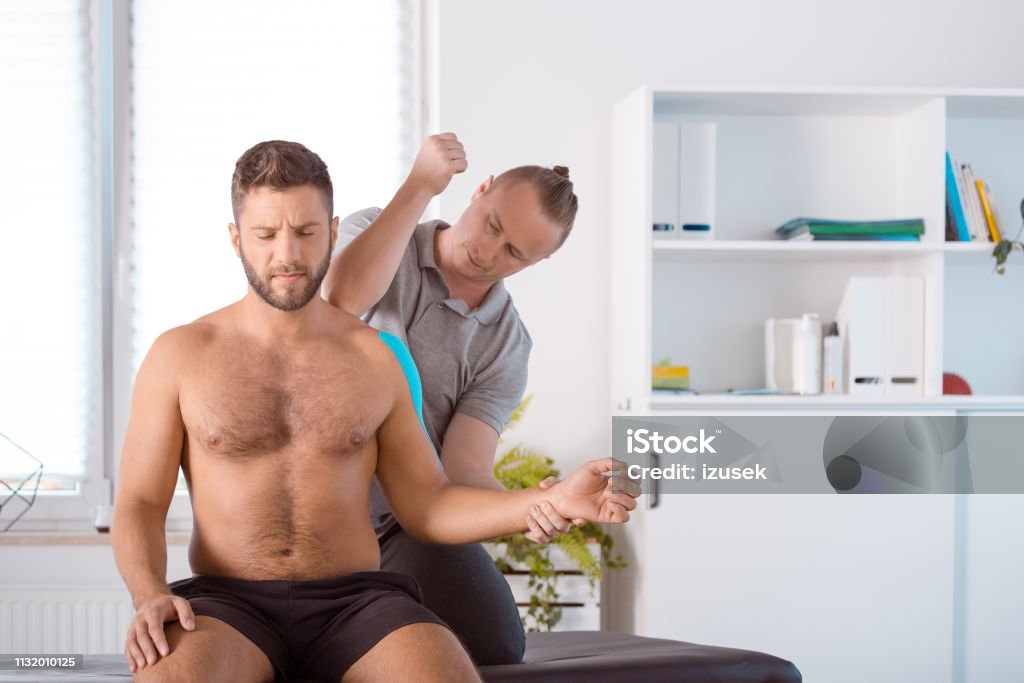 Physiotherapist massaging young man Physical therapist giving shoulder massage to young man. Patient having elastic therapeutic  tape on his arm. Adults Only Stock Photo