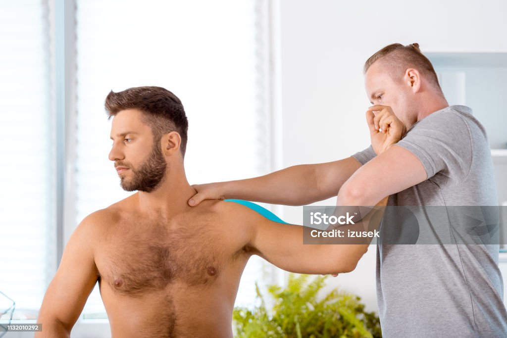 Physiotherapist stretching shoulder of young man Physical therapist giving shoulder massage to young man. Patient having elastic therapeutic  tape on his arm. Adhesive Tape Stock Photo