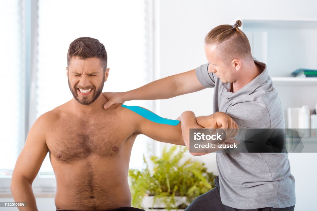 Physiotherapist stretching shoulder of young man Physical therapist giving shoulder massage to young man. Patient having elastic therapeutic  tape on his arm. Adhesive Tape Stock Photo
