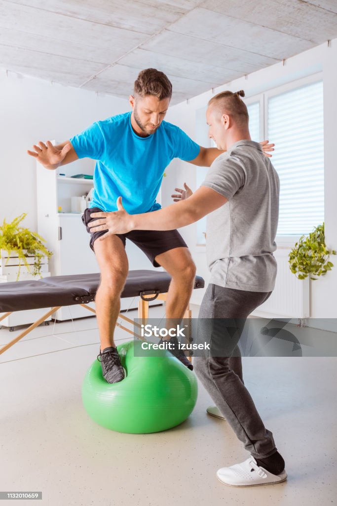 Young man excercising on ball with his physiotherapist on ball Young man having rehabilitation, standing on fitness ball and exercising with his physical therapist assistance. Balance Stock Photo