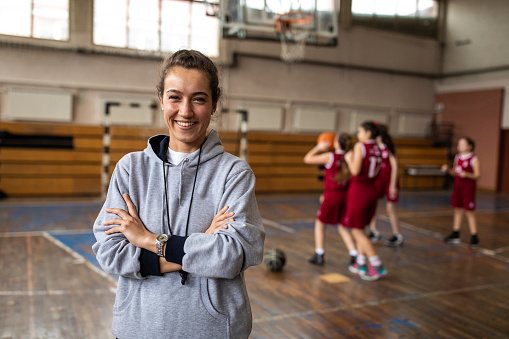 Young beautiful female basketball coach standing in school gym with crossed arms , looking at camera and smiling