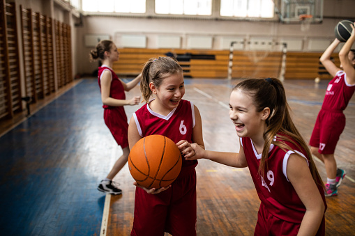 Cute teenage girls in good mood at school gym embracing and smiling on basketball field indoor