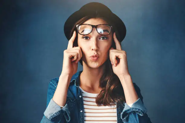 Studio shot of an attractive young woman lifting her glasses and pouting against a blue background