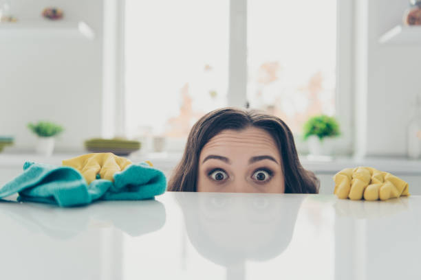 ritratto di lei lei bella bella bella bella ragazza divertente scioccato dai capelli ondulati casa-moglie che si nasconde dietro il tavolo lucido brillante in moderni interni bianchi leggeri all'interno - cleaning domestic kitchen white duster foto e immagini stock