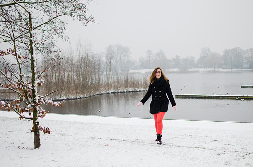 Photograph of a young girl playing and walking in a newly snowed park with a lake behind in the Netherlands.
