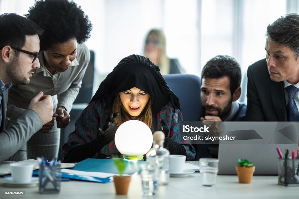 Young fortune teller predicting the future for business team. Female fortune teller analyzing crystal ball while predicting the future of business people. Crystal Ball Stock Photo