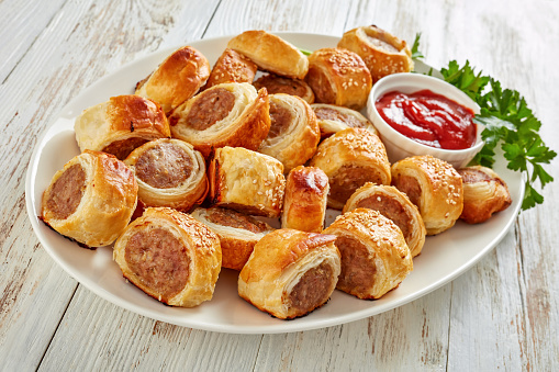 freshly baked Puff pastry Sausage rolls with tomato sauce and parsley on a white plate, english party food, horizontal view from above, close-up