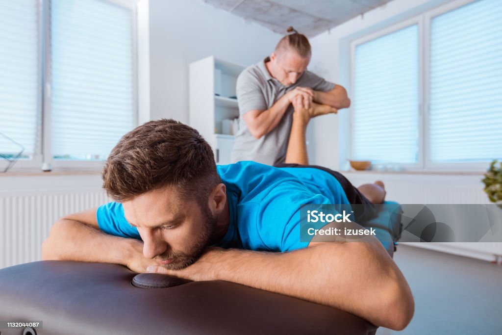 Physiotherapist massaging young man Physical therapist giving leg massage to young man. Patient lying down on massage table. Adult Stock Photo