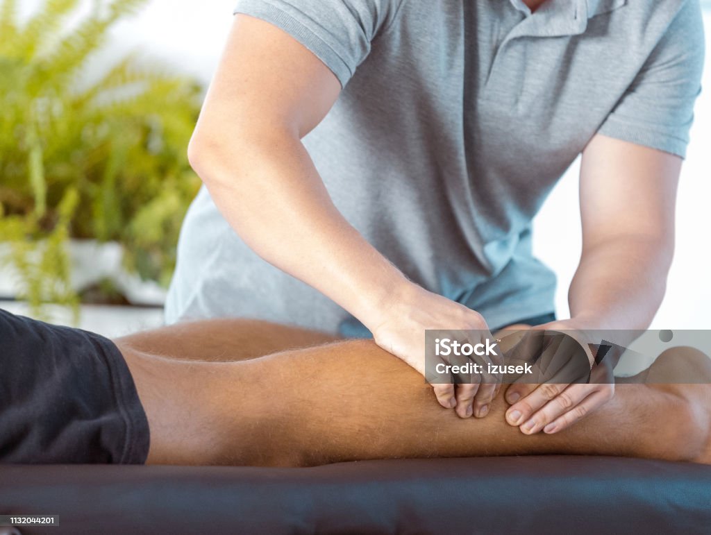 Physiotherapist massaging young man's leg Physical therapist giving leg massage to young man. Patient lying down on massage table. Close up of hands, unrecognizable person. Adult Stock Photo