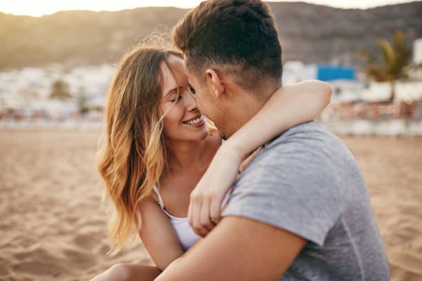 Romantic young couple sitting together on a sandy beach Young couple enjoying a romantic moment together while sitting on a sandy beach at sunset falling in love stock pictures, royalty-free photos & images